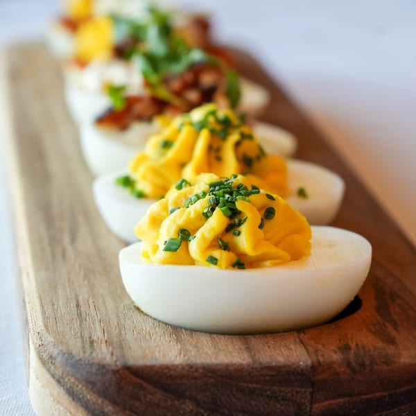 Deviled eggs with chives on a wooden board, with other garnished deviled eggs blurred in the background.