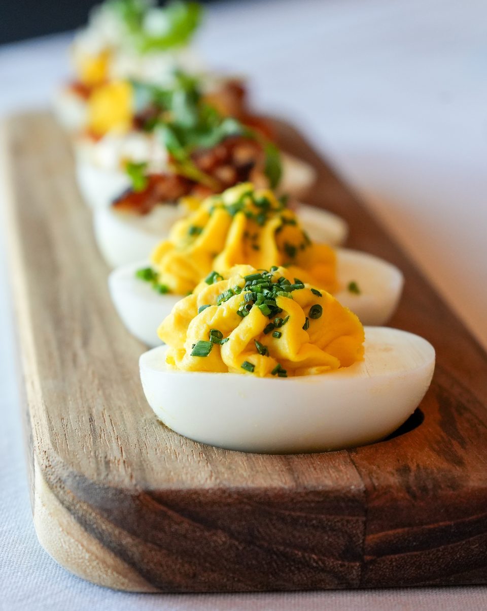Deviled eggs with chives on a wooden board, with other garnished deviled eggs blurred in the background.