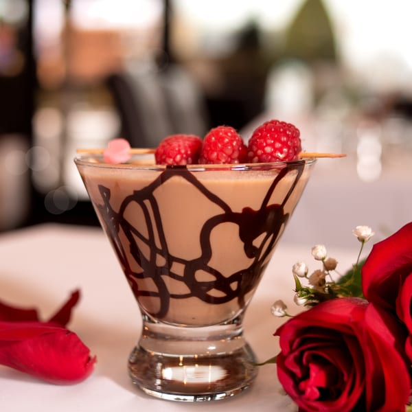 Chocolate martini with raspberries and a heart pick, alongside red roses on a white tablecloth.