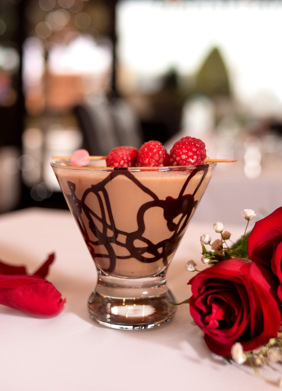Chocolate martini with raspberries and a heart pick, alongside red roses on a white tablecloth.