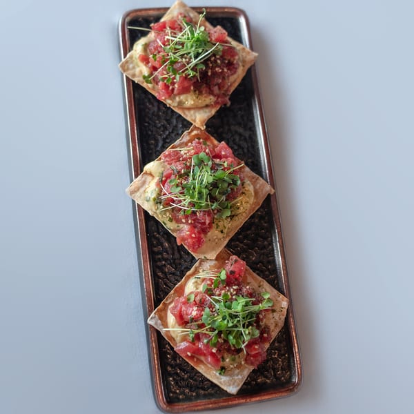 Three tuna tartare appetizers on square crackers, topped with microgreens, on a rectangular black plate.