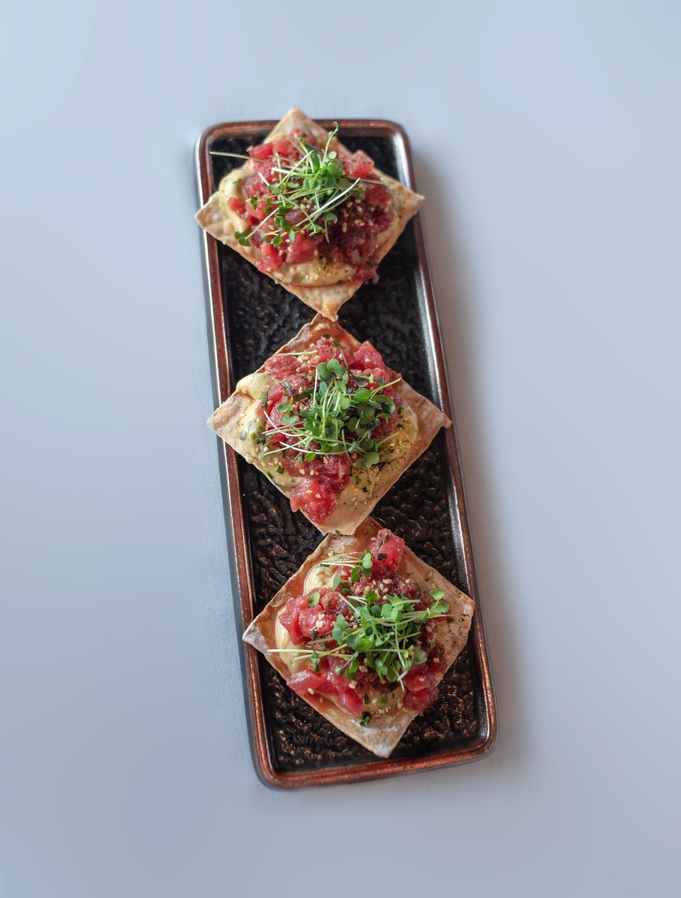 Three tuna tartare appetizers on square crackers, topped with microgreens, on a rectangular black plate.