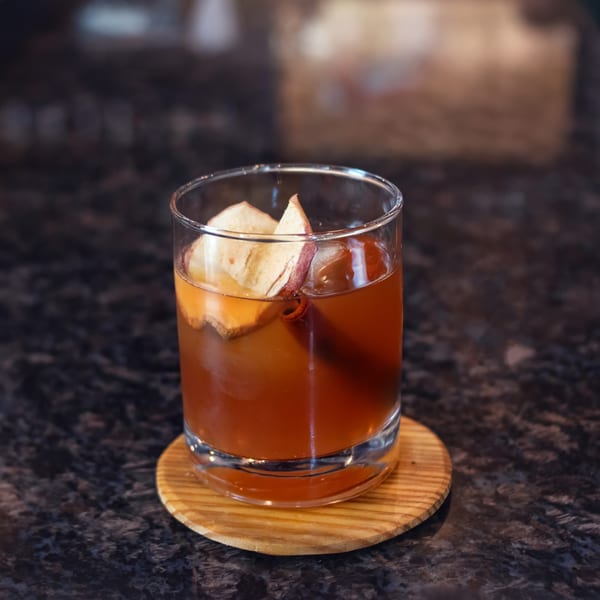 A brown cocktail with apple slices and a cinnamon stick sits on a wooden coaster.