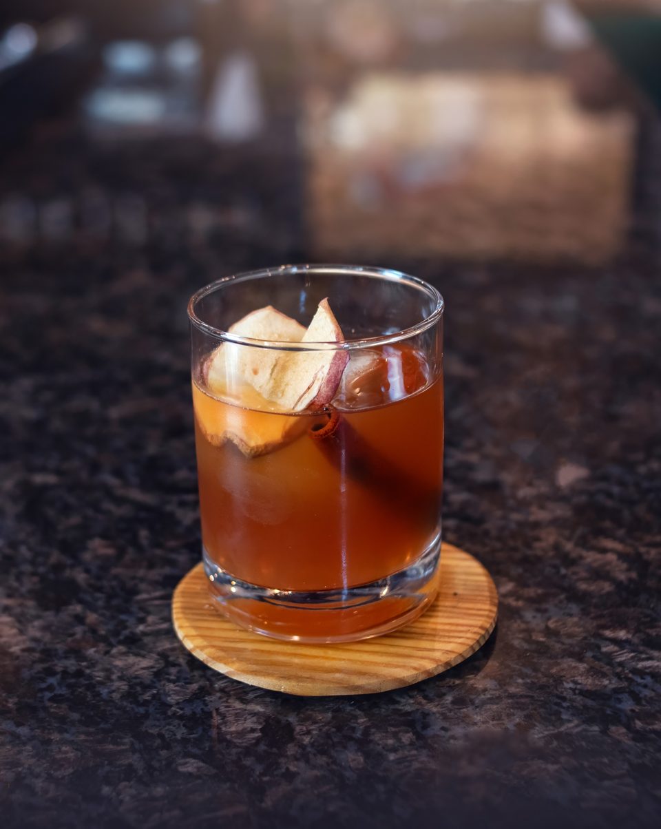 A brown cocktail with apple slices and a cinnamon stick sits on a wooden coaster.