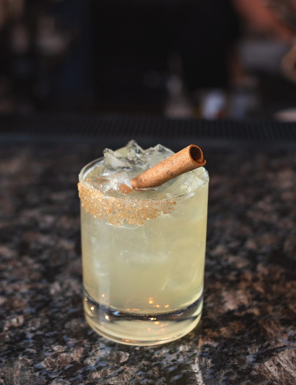 A light-colored cocktail with ice and a cinnamon stick, rimmed with brown sugar, on a dark countertop.