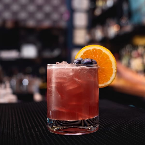 A vibrant red cocktail with ice, garnished with blueberries and an orange slice, on a dark bar.