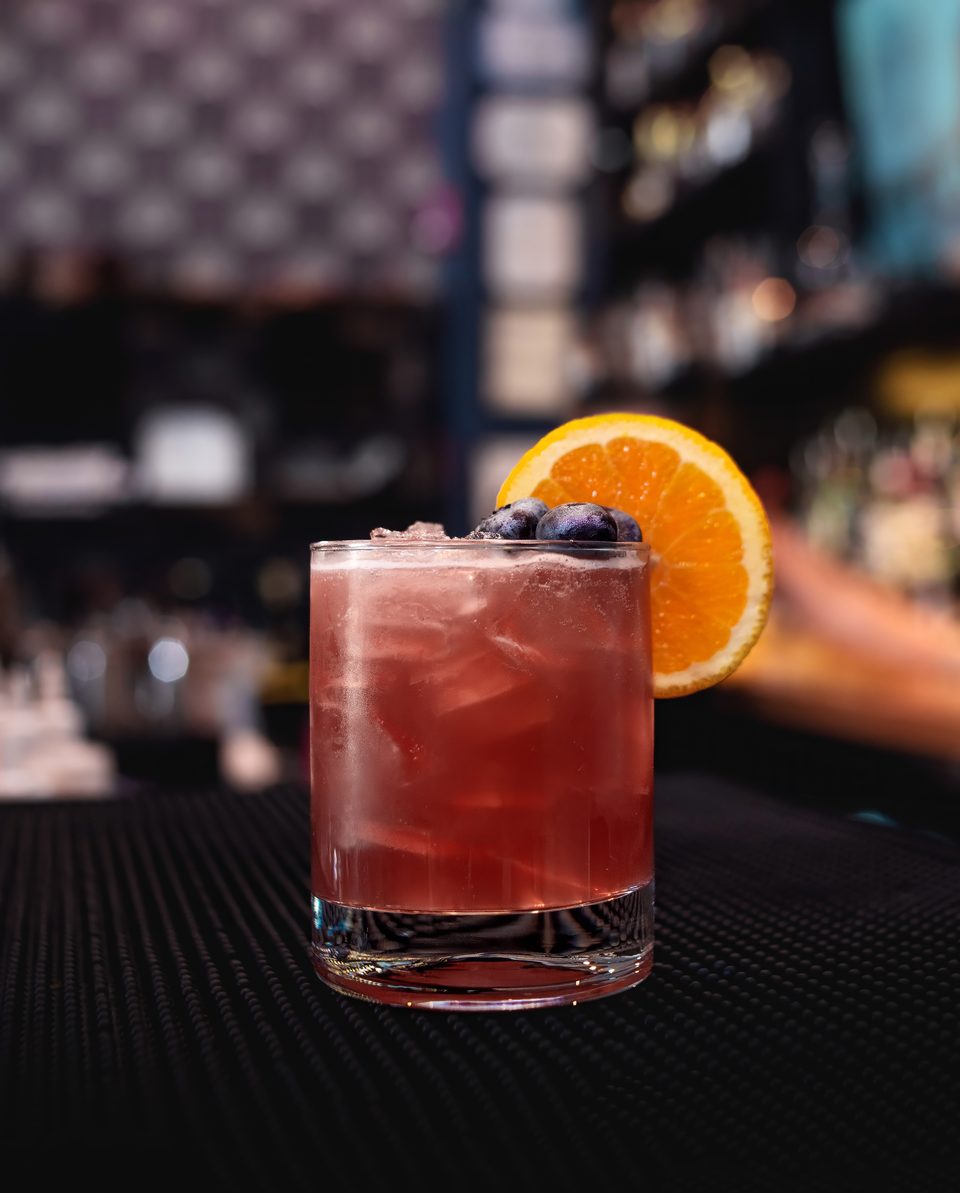 A vibrant red cocktail with ice, garnished with blueberries and an orange slice, on a dark bar.