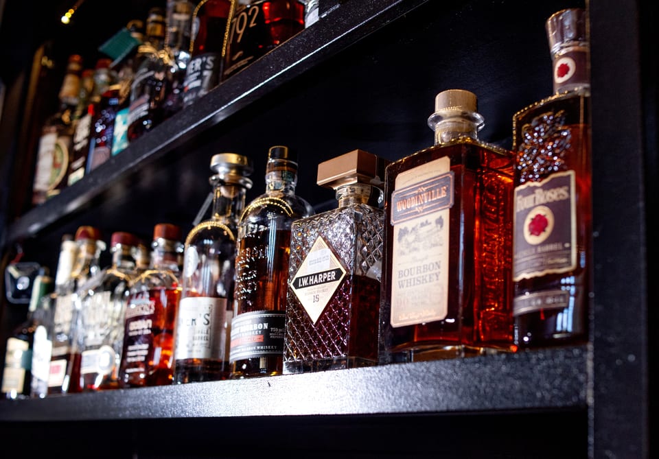 Close-up of various whiskey and bourbon bottles, including I.W. Harper and Four Roses, on a dark shelf.