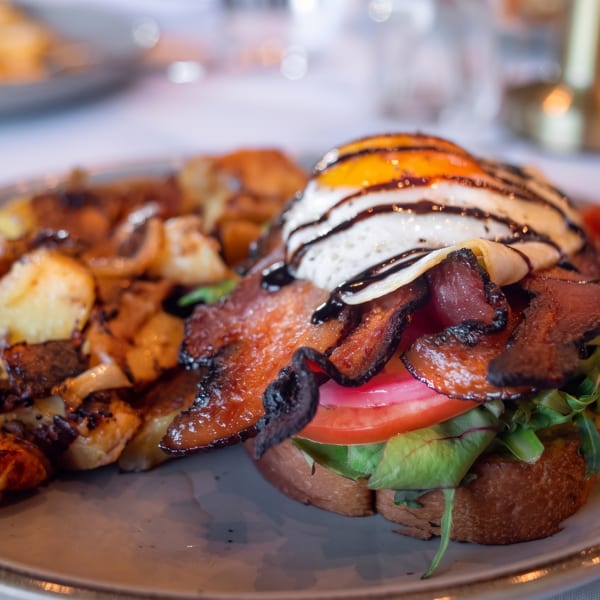 Breakfast plate with crispy bacon, fried egg, tomato, greens on toast, and roasted potatoes.