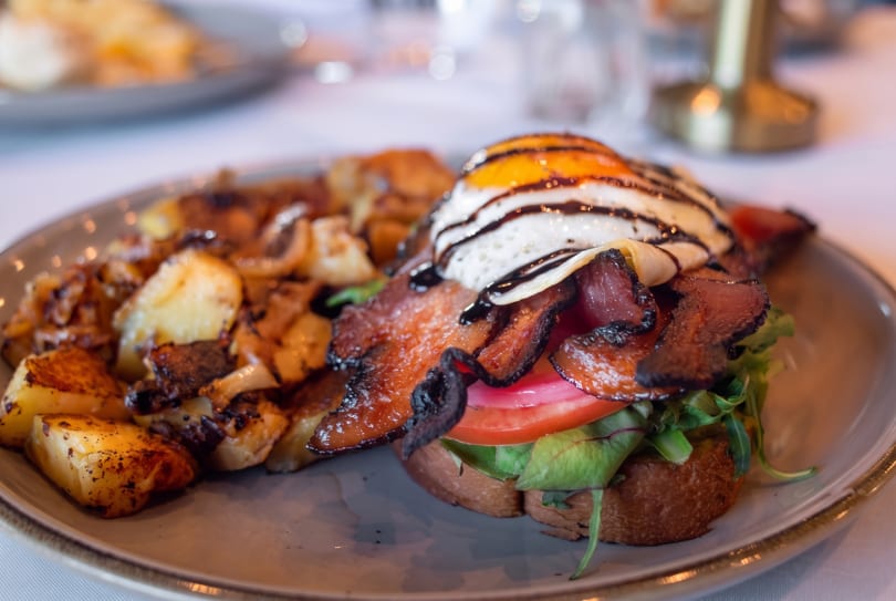 Breakfast plate with crispy bacon, fried egg, tomato, greens on toast, and roasted potatoes.