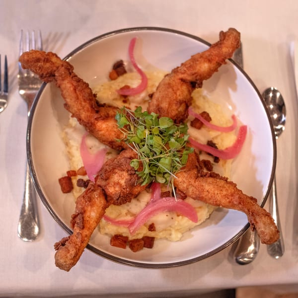 Fried frog legs with mashed potatoes, pickled onions, bacon bits, and microgreens on a white plate.