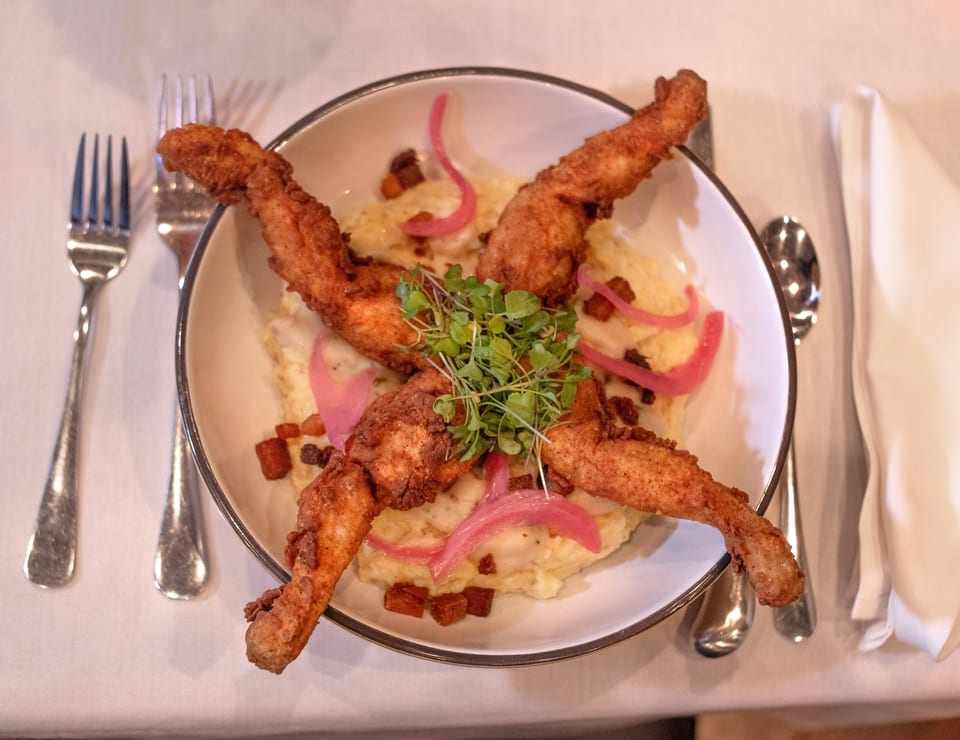 Fried frog legs with mashed potatoes, pickled onions, bacon bits, and microgreens on a white plate.