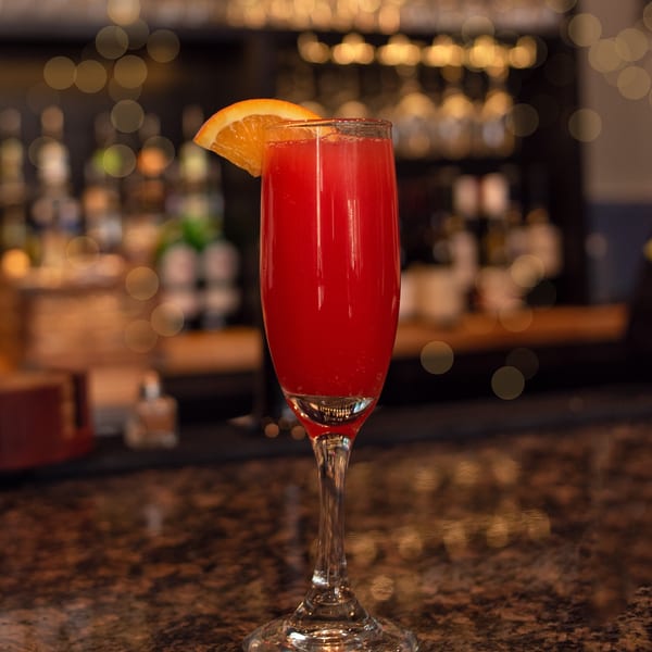 Red cocktail in a champagne flute with an orange slice, on a dark bar with bokeh lights.