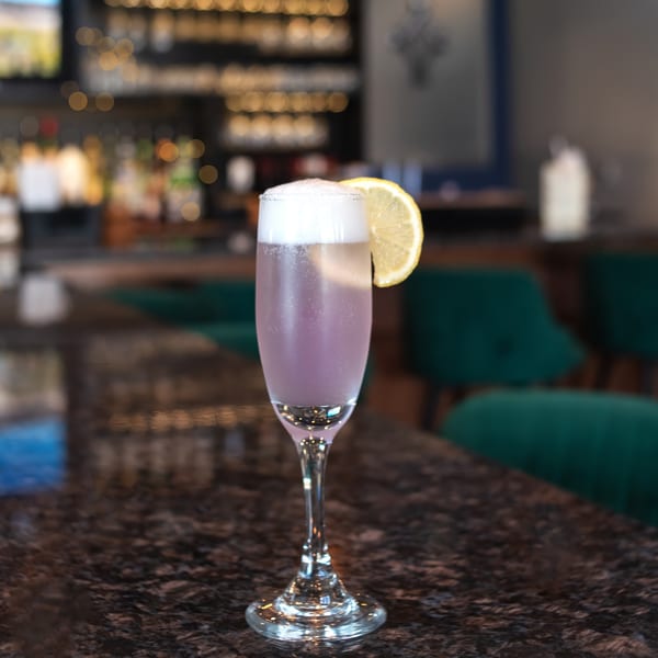 A purple cocktail with foam and a lemon slice sits on a dark bar counter.