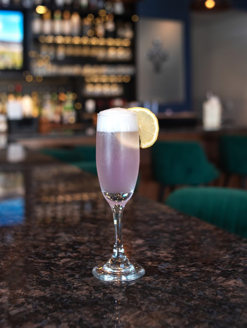 A purple cocktail with foam and a lemon slice sits on a dark bar counter.