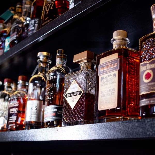 Bottles of bourbon whiskey, including I.W. Harper, Woodinville, and Four Roses, on a dark shelf.