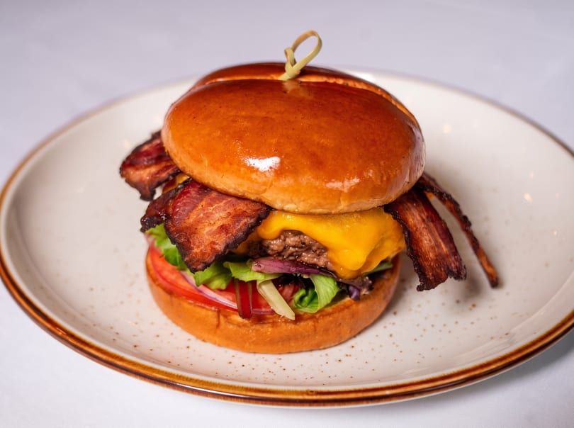 Gourmet bacon cheeseburger with lettuce, tomato, and onion on a brioche bun, served on a white plate.