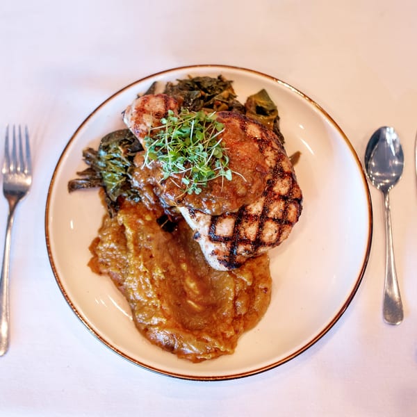 Grilled pork chop with applesauce, collard greens, and microgreens on a white plate with cutlery.