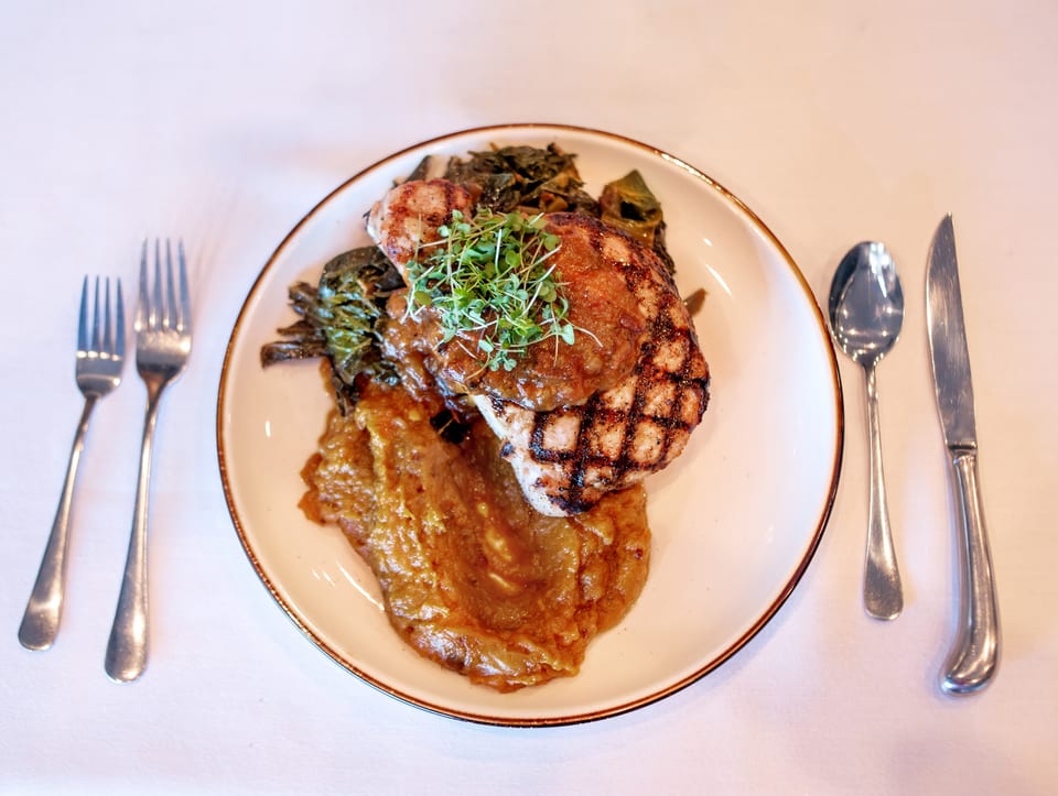 Grilled pork chop with applesauce, collard greens, and microgreens on a white plate with cutlery.