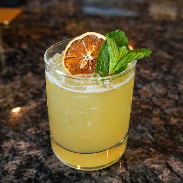A yellow cocktail with ice, garnished with a dried orange slice and fresh mint, on a dark granite surface.