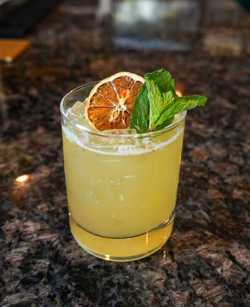 A yellow cocktail with ice, garnished with a dried orange slice and fresh mint, on a dark granite surface.