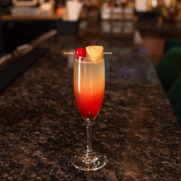 A vibrant red and orange cocktail, garnished with pineapple and a cherry, sits on a dark granite bar.