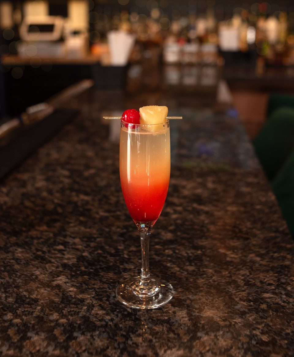 A vibrant red and orange cocktail, garnished with pineapple and a cherry, sits on a dark granite bar.
