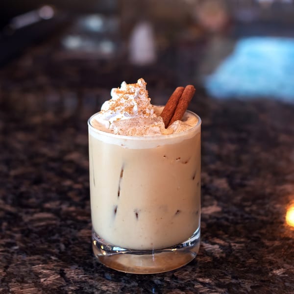 Creamy cocktail with whipped cream, cinnamon sticks, and spice on a dark granite bar.