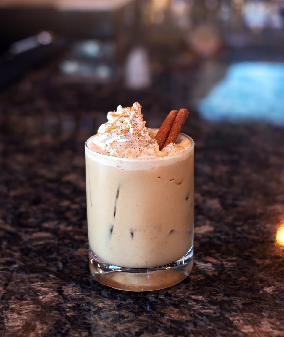 Creamy cocktail with whipped cream, cinnamon sticks, and spice on a dark granite bar.