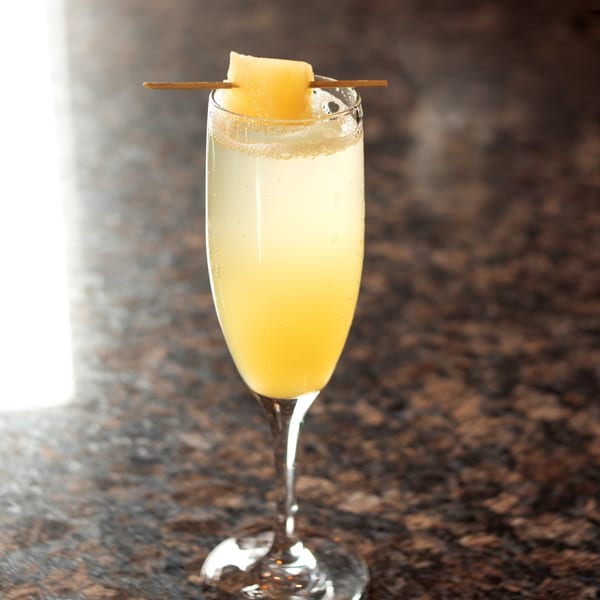Sparkling yellow cocktail in a champagne flute, garnished with a cantaloupe cube on a skewer.