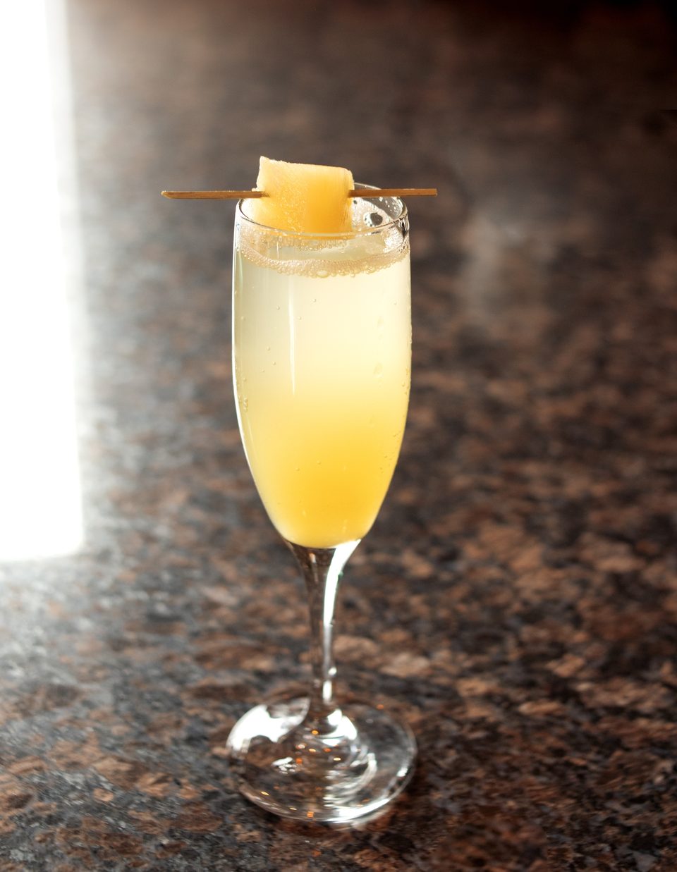 Sparkling yellow cocktail in a champagne flute, garnished with a cantaloupe cube on a skewer.