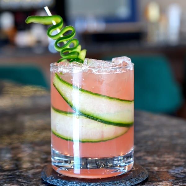Pink cocktail with cucumber slices and a cucumber spiral garnish on a dark marble bar.