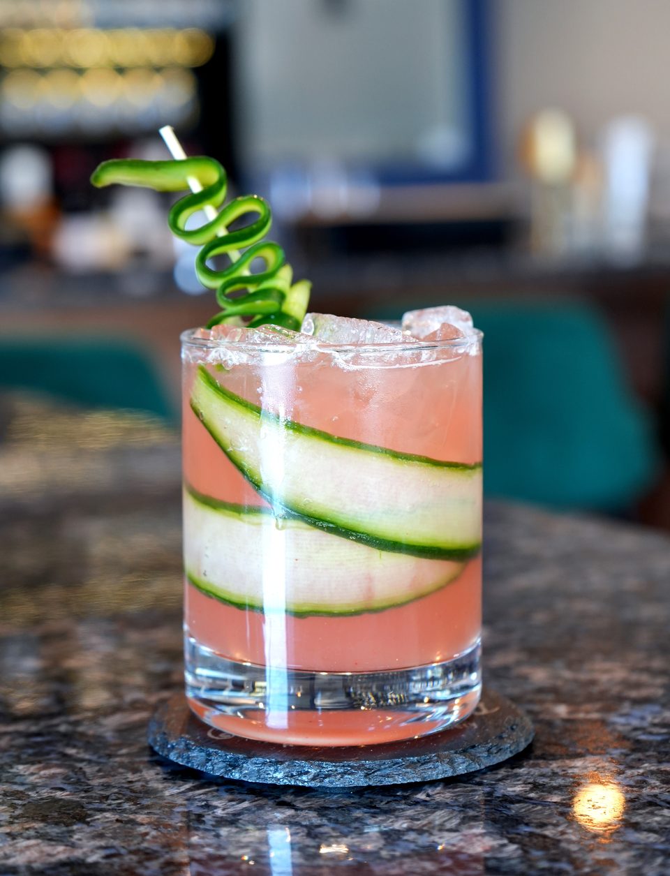 Pink cocktail with cucumber slices and a cucumber spiral garnish on a dark marble bar.