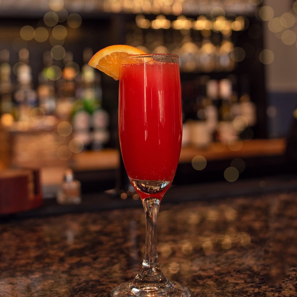 A red cocktail with an orange slice garnish sits on a dark bar with bokeh lights in the background.