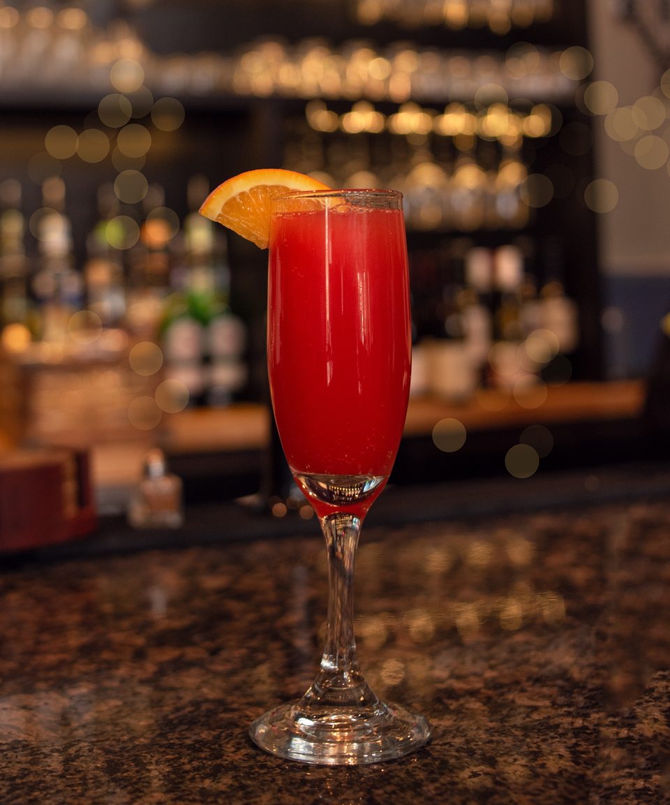 A red cocktail with an orange slice garnish sits on a dark bar with bokeh lights in the background.