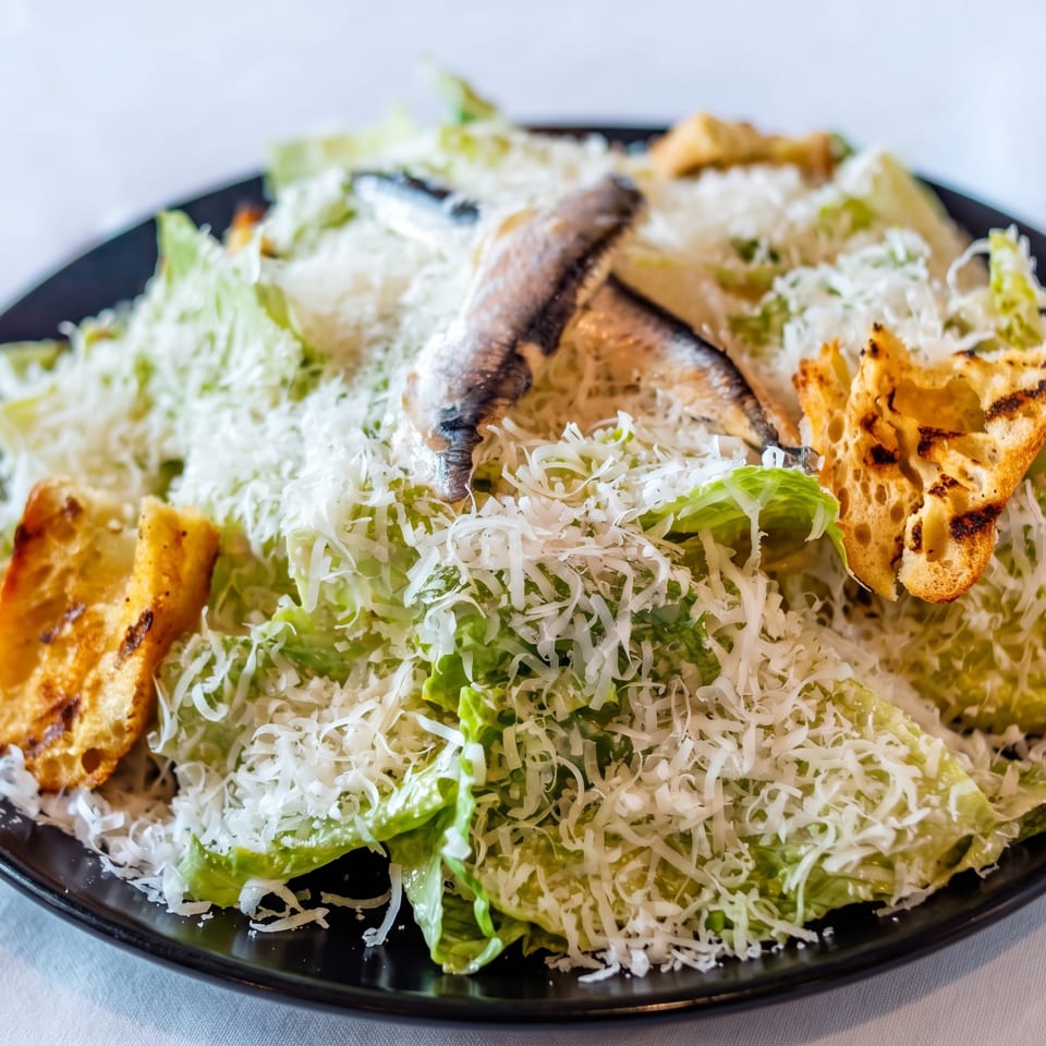 Caesar salad with anchovies, croutons, and shredded Parmesan cheese on a black plate.