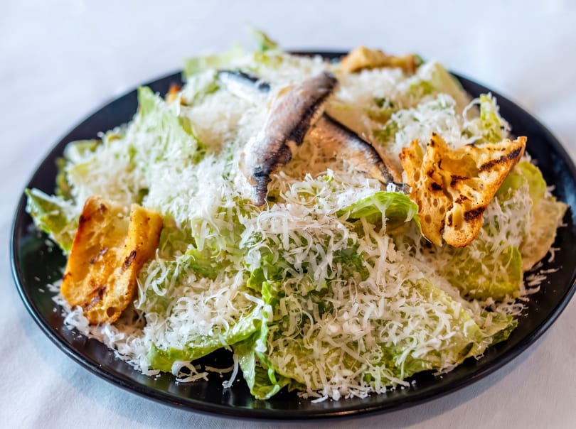 Caesar salad with anchovies, croutons, and shredded Parmesan cheese on a black plate.