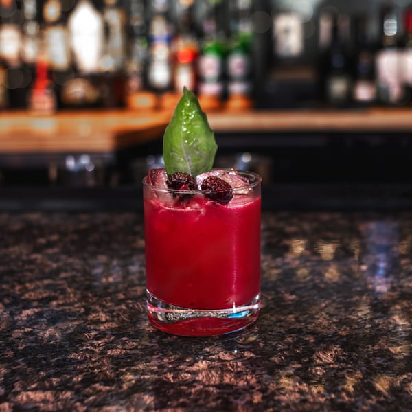 A vibrant red cocktail with ice, garnished with blackberries and a green leaf, on a dark bar.