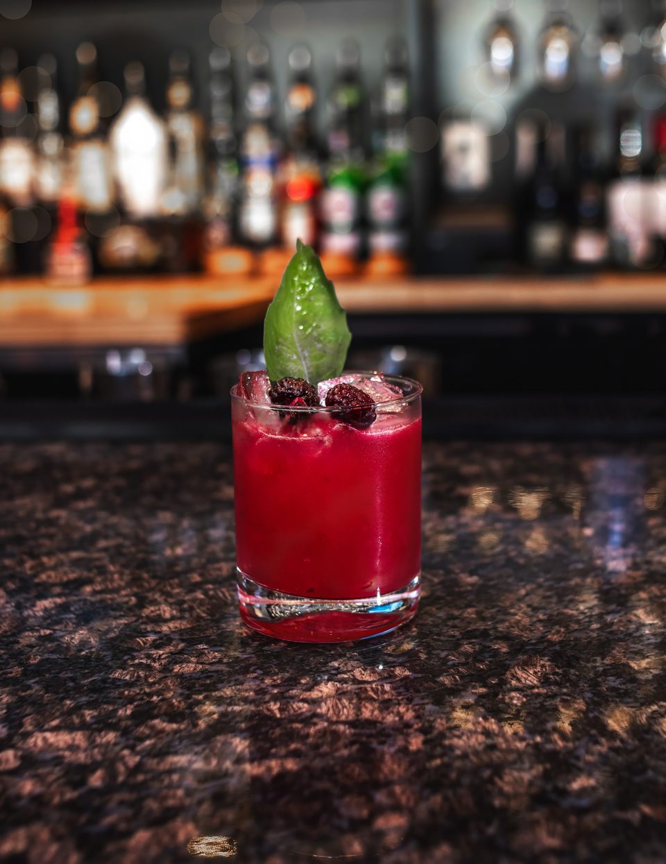 A vibrant red cocktail with ice, garnished with blackberries and a green leaf, on a dark bar.