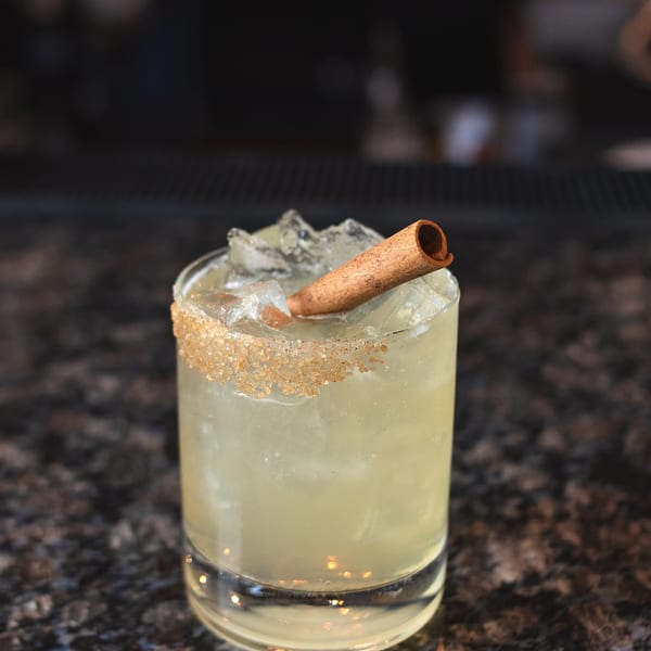 A light-colored cocktail with ice, a cinnamon stick, and a sugar-rimmed glass on a dark countertop.
