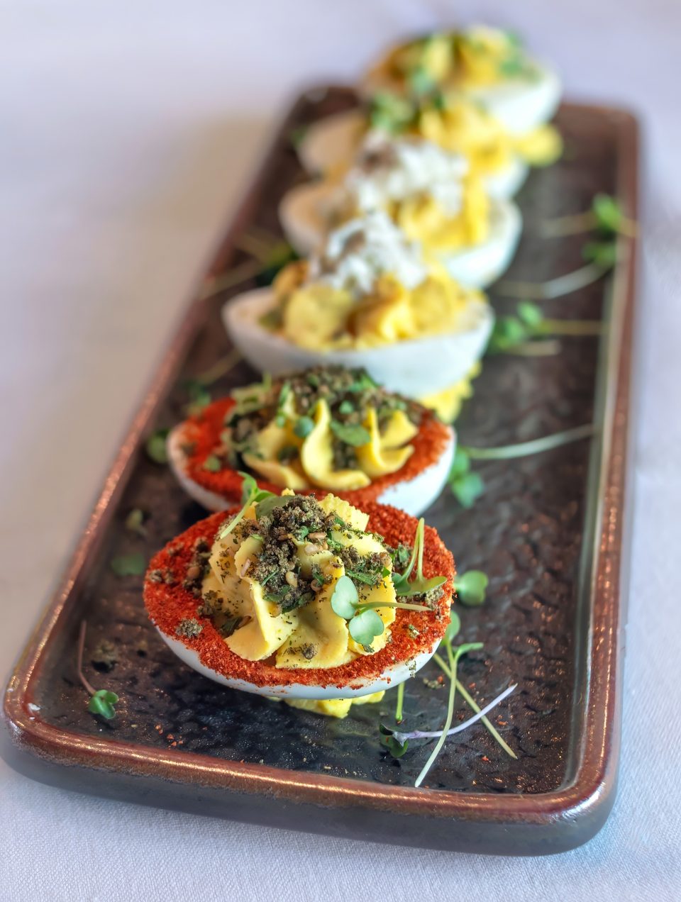 A rectangular platter holds five deviled eggs, two with red paprika and green herbs, three with white topping.